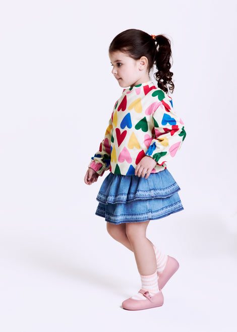 Child model in a heart shirt and blue skirt wearing a pair of milky pink Soft Ballerina baby flats