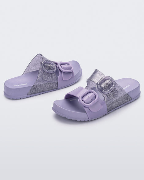 An angled front and side view of a pair of lilac Mini Melissa Cozy slides with two front straps with buckle details