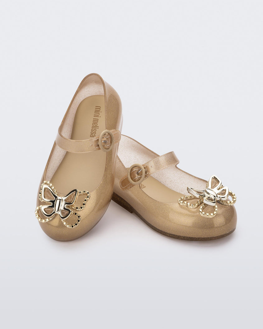 An angled front and top view of a pair of glitter beige Mini Melissa Sweet Love Butterfly flats, leaning on eachother, with a top strap and a gold butterfly detail on the toe