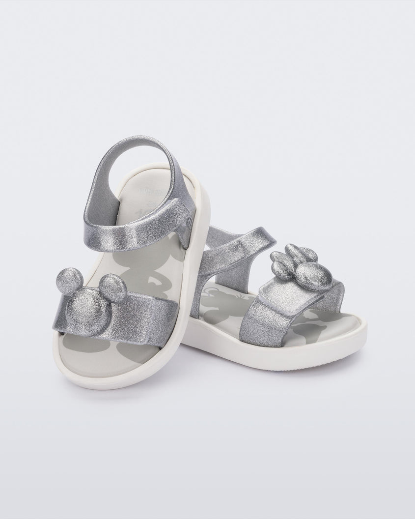An angled top and side view of a pair of white/glitter silver Mini Melissa Jump sandals, leaning on eachother, with a Mickey Mouse logo detail on the front strap and an ankle strap