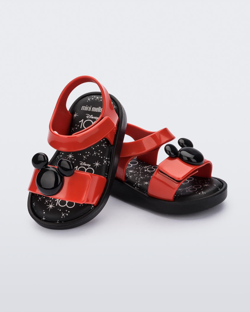 An angled top and side view of a pair of red/black Mini Melissa Jump sandals, leaning on eachother, with a black Mickey Mouse logo detail on the front red strap and a red ankle strap