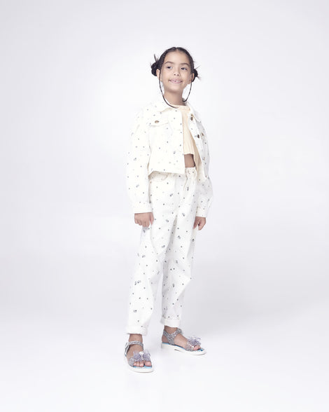 A kid model posing for a picture in a white patterned top and bottom, wearing a pair of glitter clear Mini Melissa Mar Sandal Princess sandals with a snowflake bow detail on the front strap, an ankle strap and Princess Elsa soul