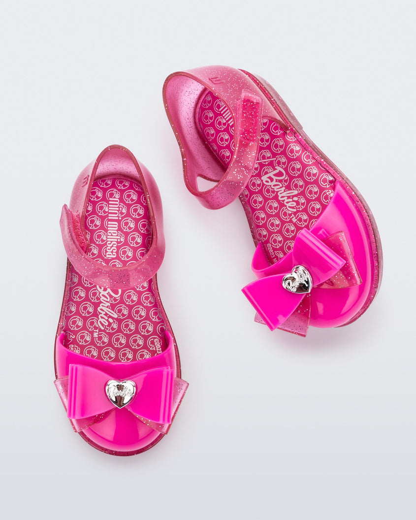 A top view of a pair of glitter pink Mini Melissa sandals with a Barbie bow detail on the front toe, pink glitter ankle strap and a Barbie logo sole
