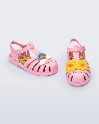 Angled view of a matte pink kids Possession + Play Doh sandal with one shoe having a daisy and the other a rainbow on the upper