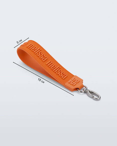 Angled flat view of an orange M Lover Charm with the Melissa logo with measurements of 15cm x 3cm