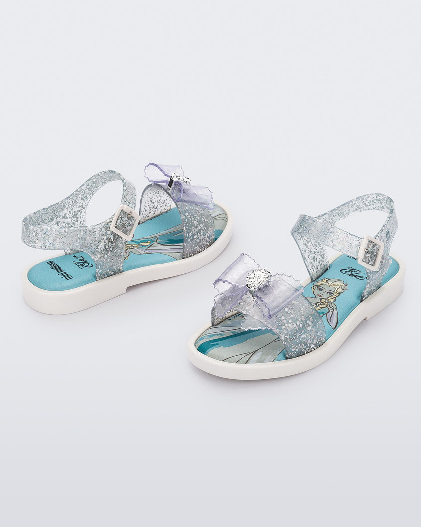 An angled front and side view of a pair of glitter clear Mini Melissa Mar Sandal Princess sandals with a snowflake bow detail on the front strap, an ankle strap and Princess Elsa soul