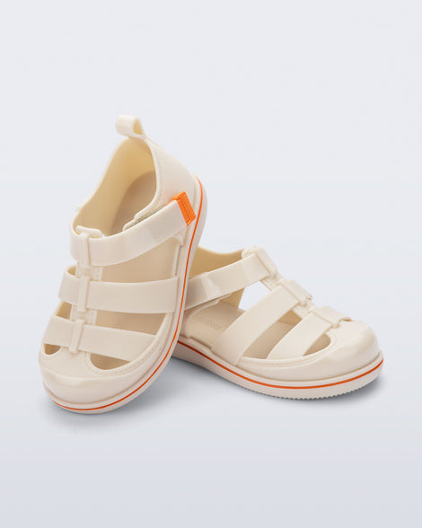 Angled view of a pair of beige Smash closed toe baby sandals with orange trim