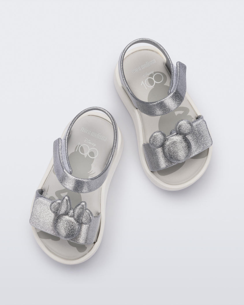 A top view of a pair of white/glitter silver Mini Melissa Jump sandals with a Mickey Mouse logo detail on the front strap and an ankle strap