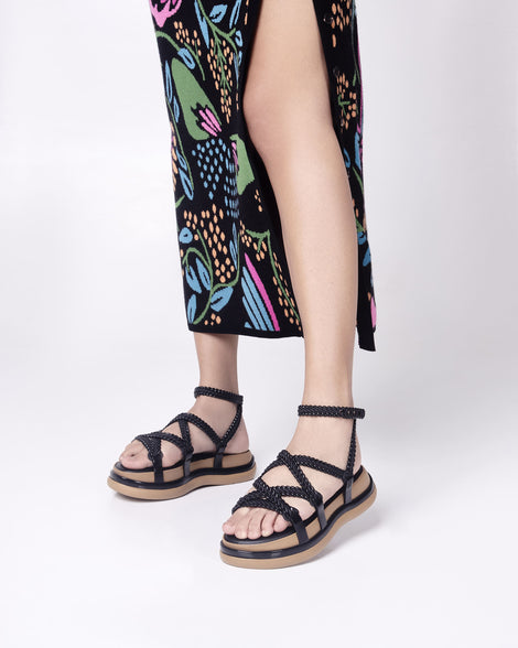 A model's legs in a patterned skirt, wearing a pair of black Melissa Buzios platform sandals with multiple textured straps that mimic sisal braids across the front of the shoe as well as an ankle strap