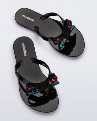 Top view of a pair black Harmonic M Lover kids flip flops with rainbow Melissa logo on bow