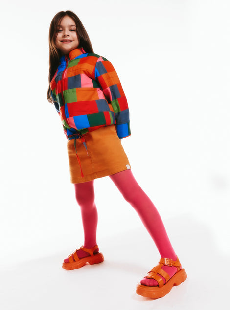 Model in an orange skirt and multicolor jacket wearing a pair of orange Ella kids sandals with pink tights