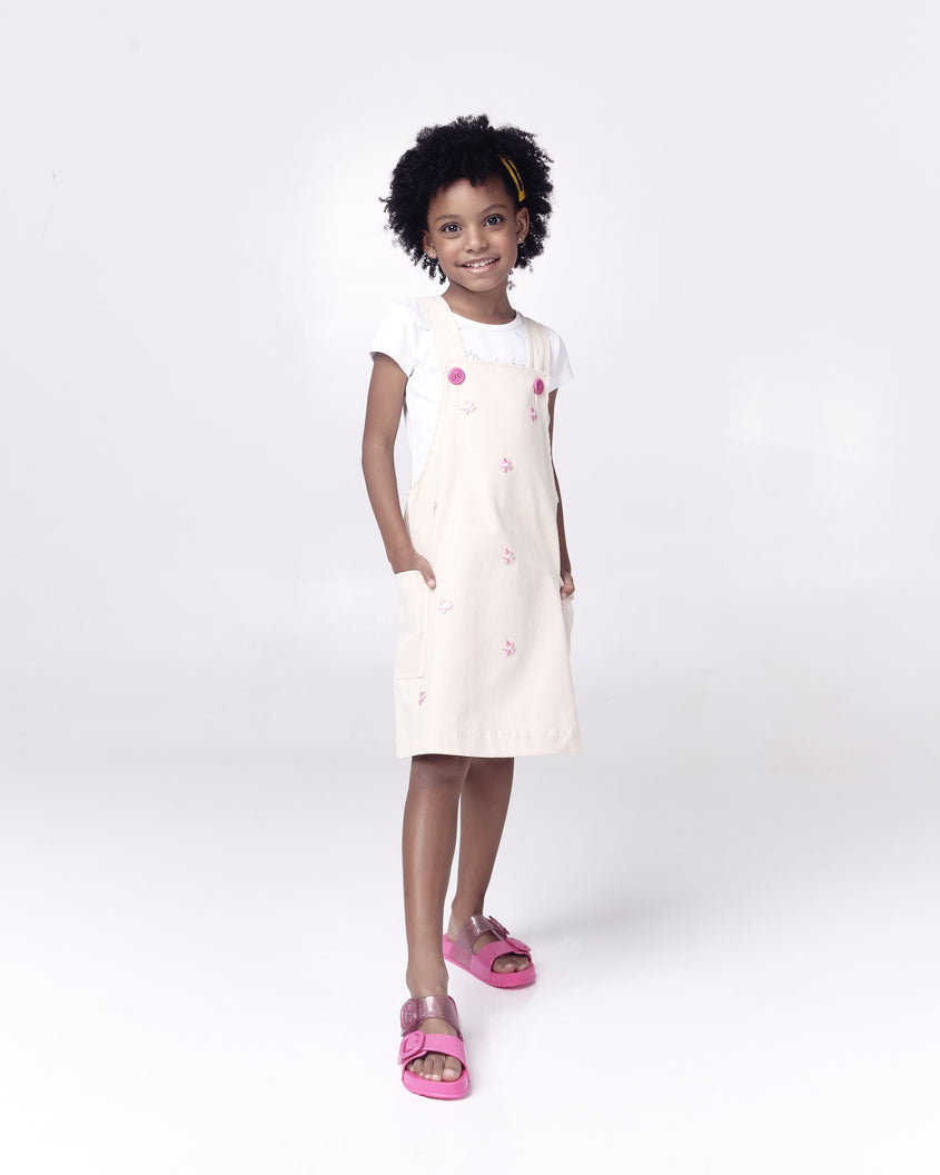 A kid model posing for a picture in a white dress, wearing a pair of pink Mini Melissa Cozy slides with two front straps with buckle details