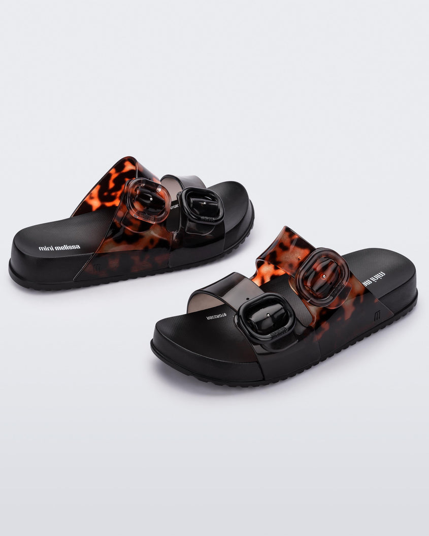 An angled front and side view of a pair of black tortoiseshell Mini Melissa Cozy slides with two front straps with buckle details