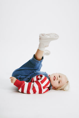 Child model in a red and white strip shirt and jean overalls wearing a pair of pearly beige Chelsea II baby boots