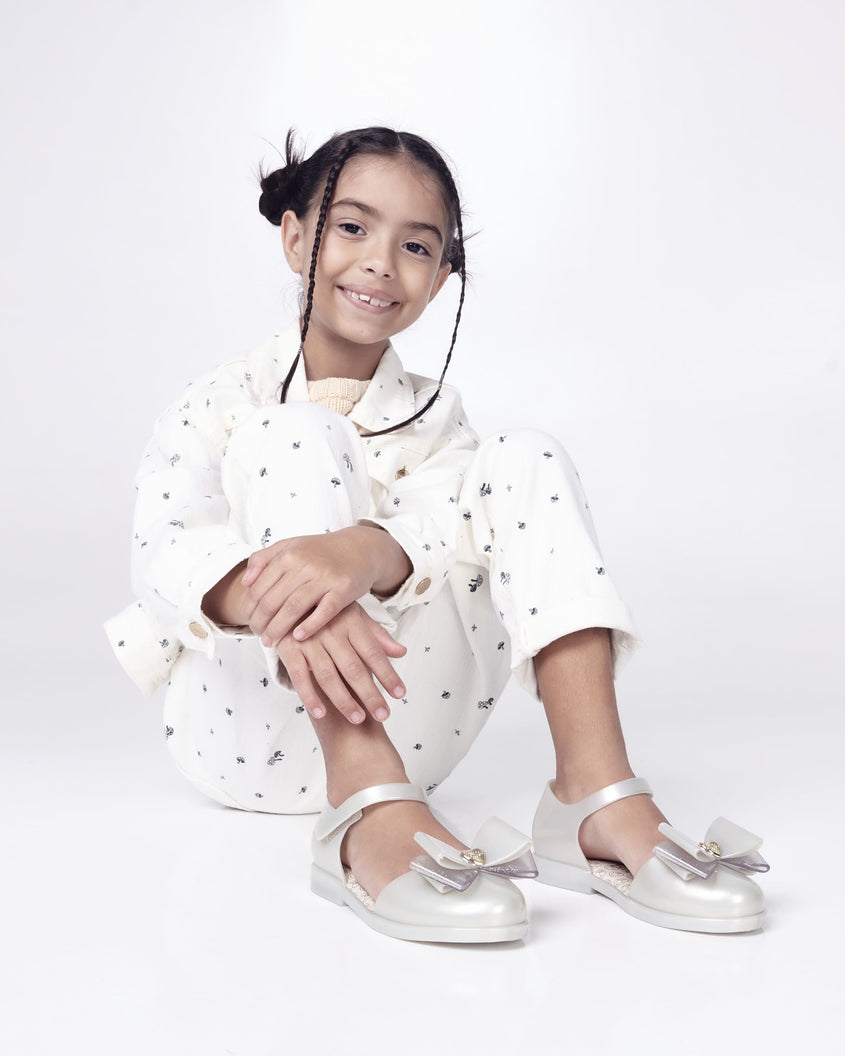 A kid model posing for a picture in a white patterned top and bottom, wearing a pair of metallic white Mini Melissa sandals with a Barbie bow detail on the front toe, metallic white ankle strap and a Barbie logo sole