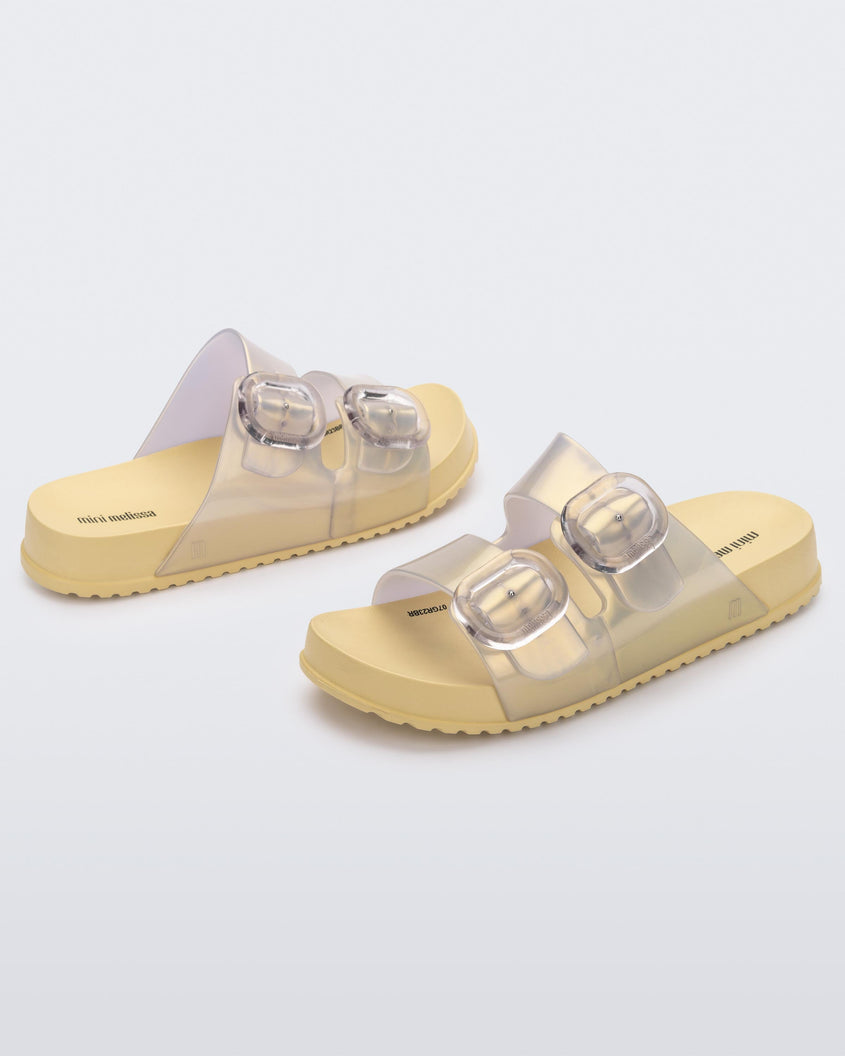 An angled front and side view of a pair of pearly yellow Mini Melissa Cozy slides with two front straps with buckle details