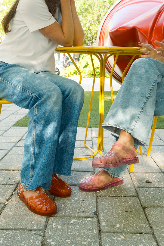 Two people sitting at a yellow metal table outdoors on stone pavers, wearing clear jelly sandals. One person wears orange Possession sandals  and the other wears translucent pinkish-brown jelly Possession sandals.