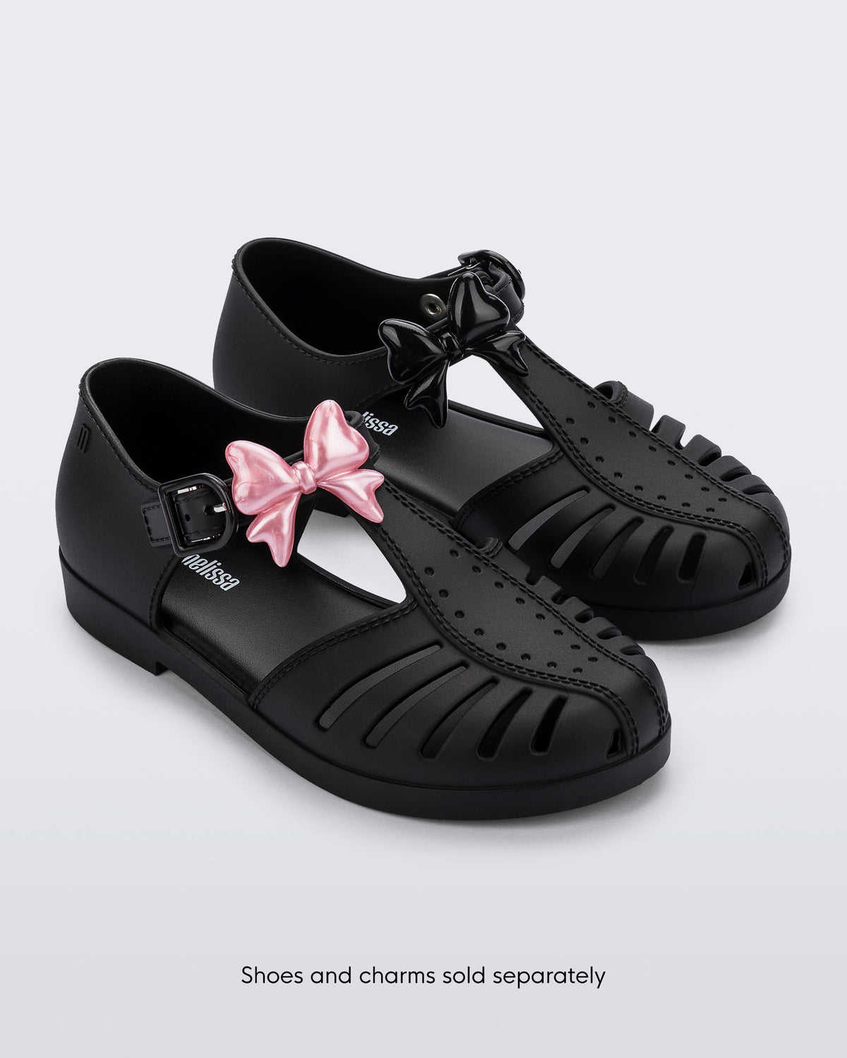 A pair of black Aranha sandals with a pearly pink bow Icons charms on the left shoe strap and black bow Icons charms on the right shoe strap. The words, Shoes and Charms sold separately on the bottom of the image