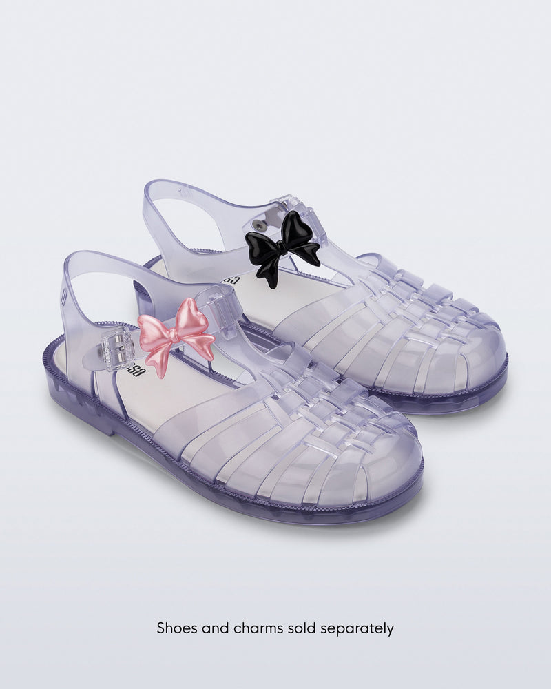 A pair of clear possession fisherman sandals with a pearly pink bow Icons charms on the left shoe strap and black bow Icons charms on the right shoe strap. The words, Shoes and Charms sold separately on the bottom of the image