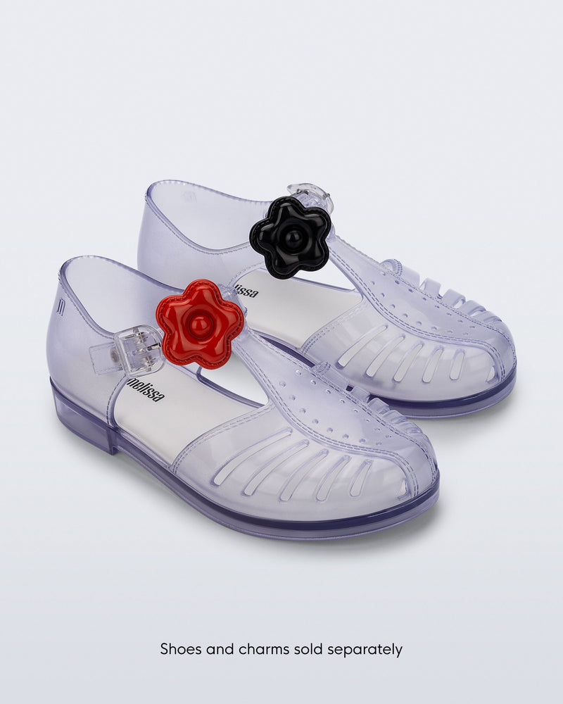 A pair of clear Aranha sandals with a red flower Icons charms on the left shoe strap and black flower Icons charms on the right shoe strap. The words, Shoes and Charms sold separately on the bottom of the imageseparately 