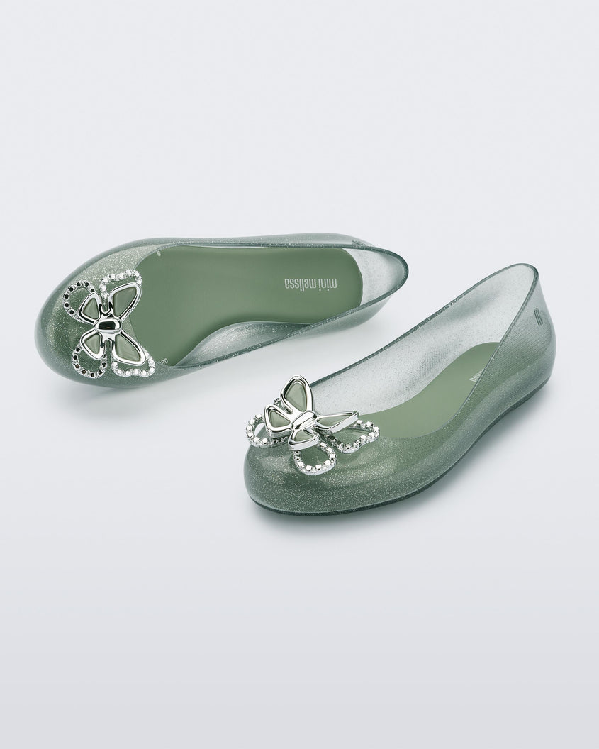 An angled front and top view of a pair of glitter green Mini Melissa Sweet Love Butterfly flats with a silver butterfly detail on the toe