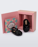 Top and angled view of a pair of black baby My First Mini Melissa Possession sandals in front of collective shoe box and book.