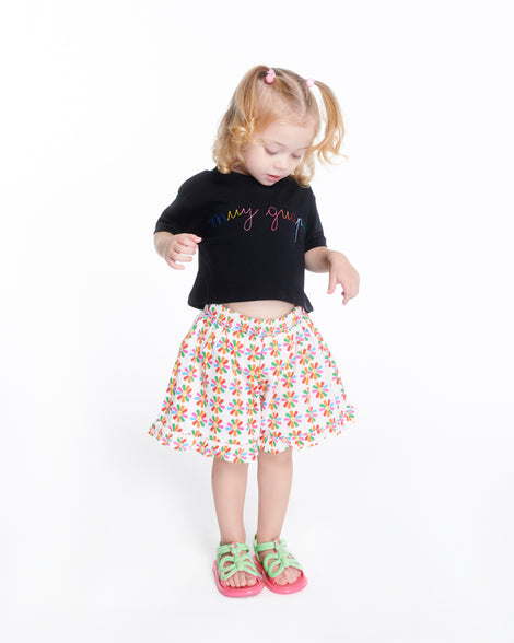 Child model in a skirt and black top wearing a pair of green strap, pink sole Hip Butterfly baby sandals