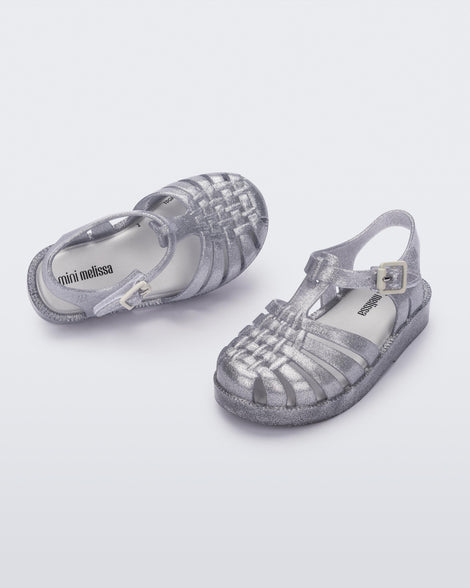 Angled view of a pair of Mini Melissa Possession baby sandals in glitter clear with velcro buckle closure on the ankle straps