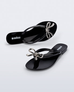 Top and angled view of a pair of black Harmonic Iris flip flops with silver bow