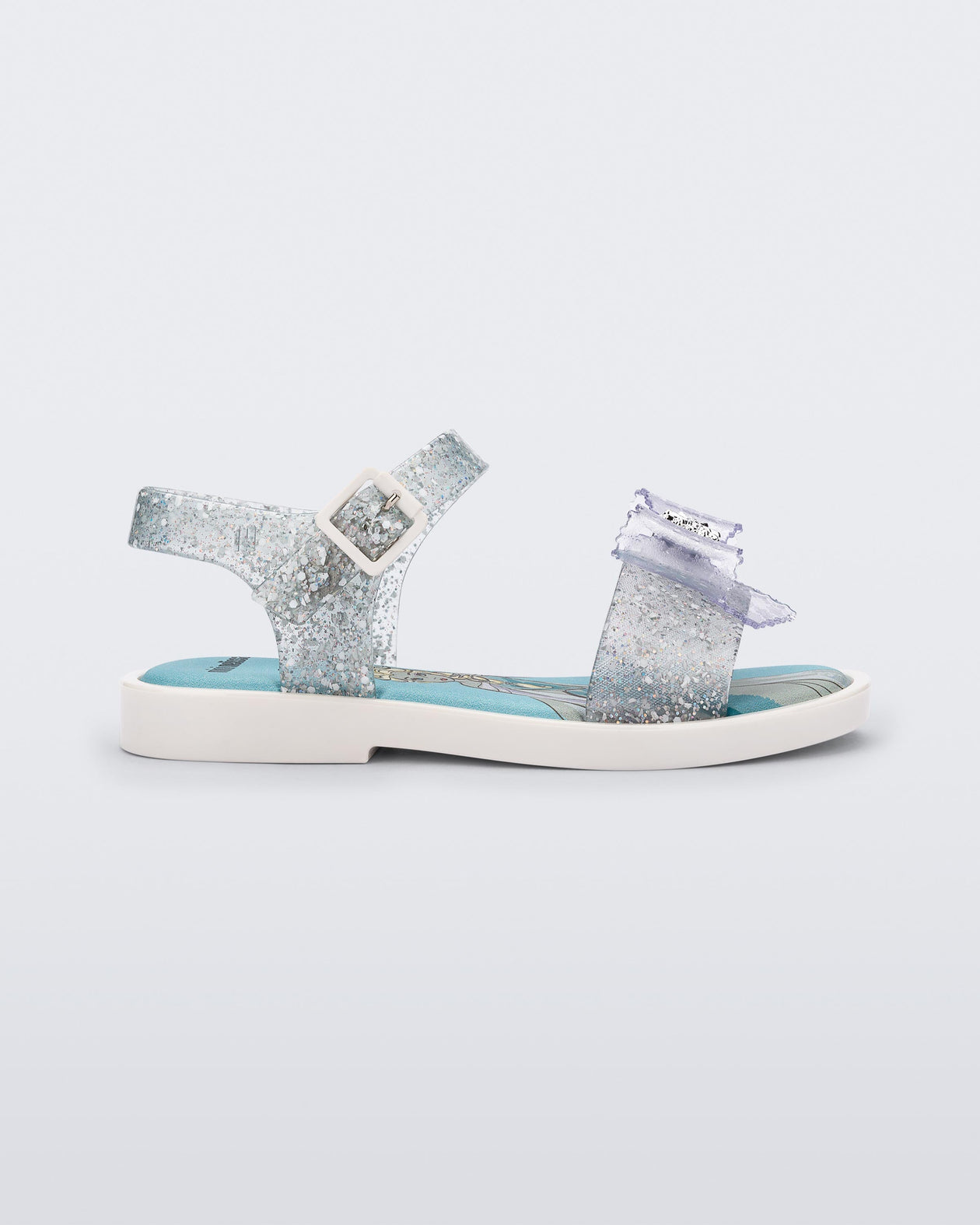 Side view of a glitter clear Mini Melissa Mar Sandal Princess sandal with a snowflake bow detail on the front strap, an ankle strap and Princess Elsa soul