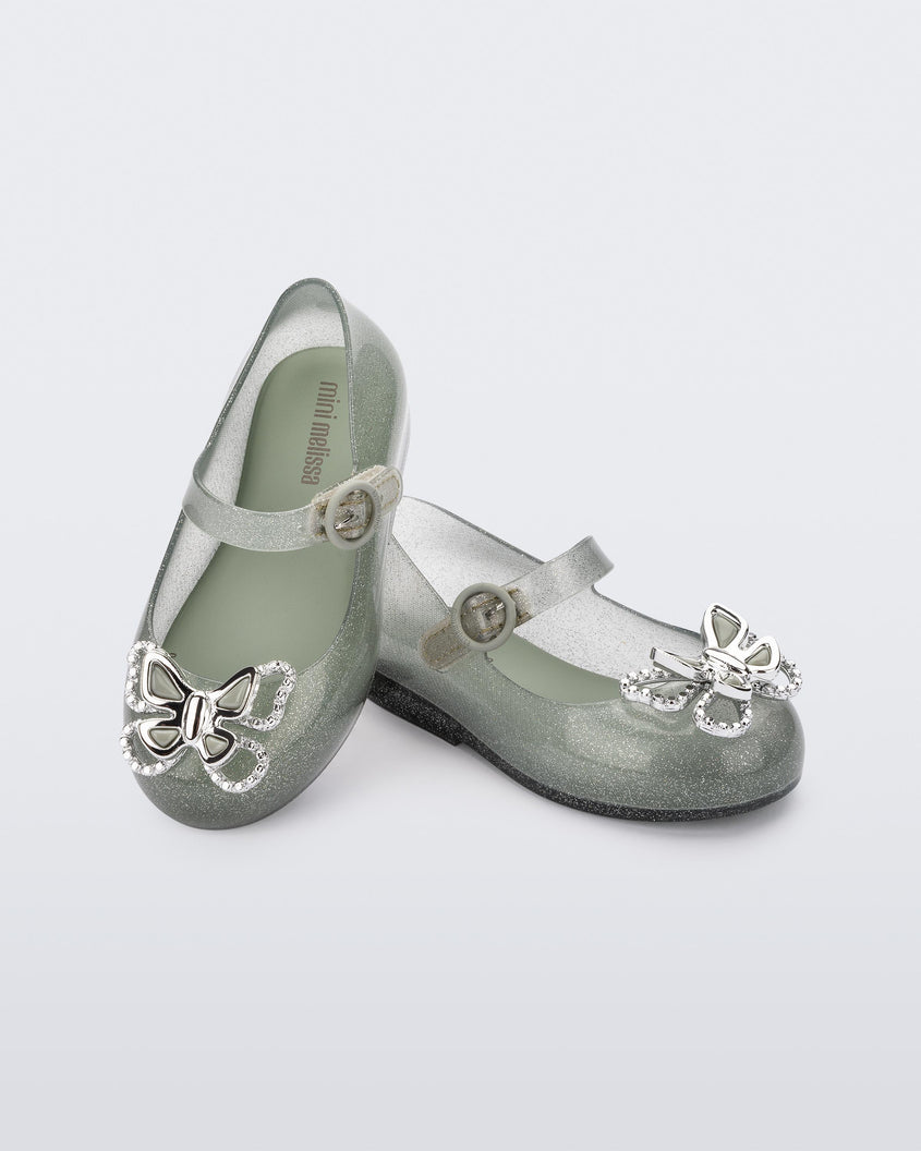 An angled front and top view of a pair of glitter green Mini Melissa Sweet Love Butterfly flats, leaning on eachother, with a top strap and a silver butterfly detail on the toe