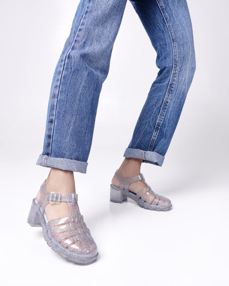 Model's legs in a pair of jeans wearing a pair of clear glitter Possession Heel women's fisherman style sandals.