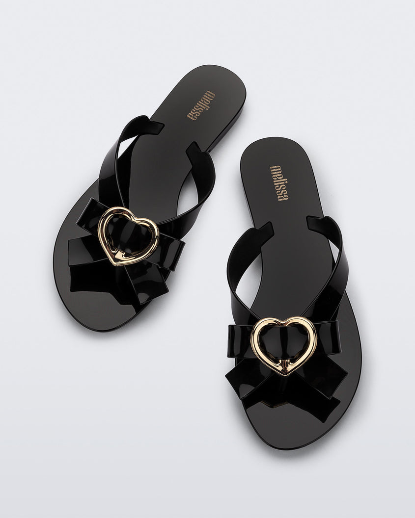 Top view of a pair of black Melissa Harmonic Heart flip flops with a black bow and gold heart detail on the straps