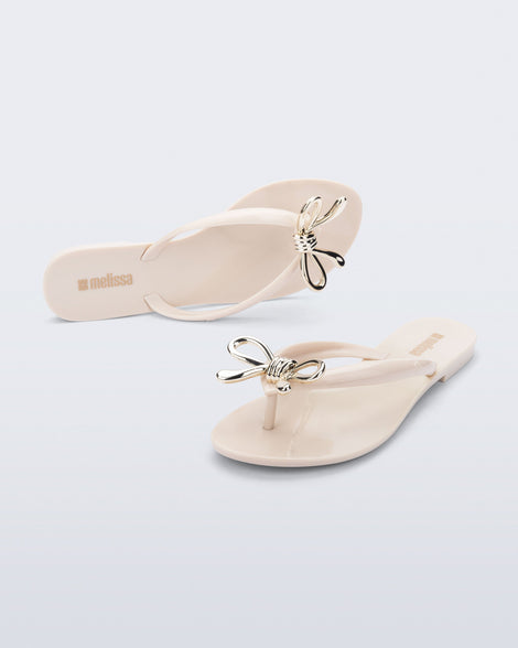 Top and angled view of a pair of beige Harmonic Iris flip flops with gold bow