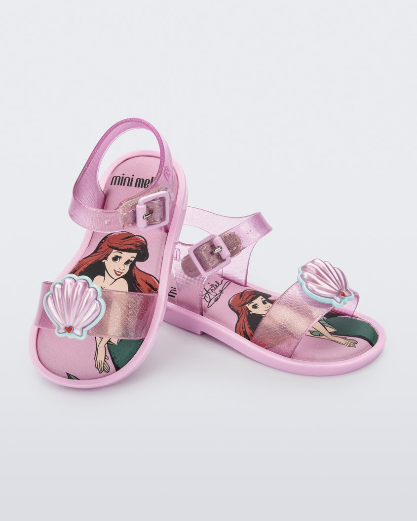 An angled top and side view of a pair of glitter pink Mini Melissa Mar Sandal Princess sandals, leaning on eachother, with a seashell detail on the front strap, an ankle strap and Princess Ariel soul