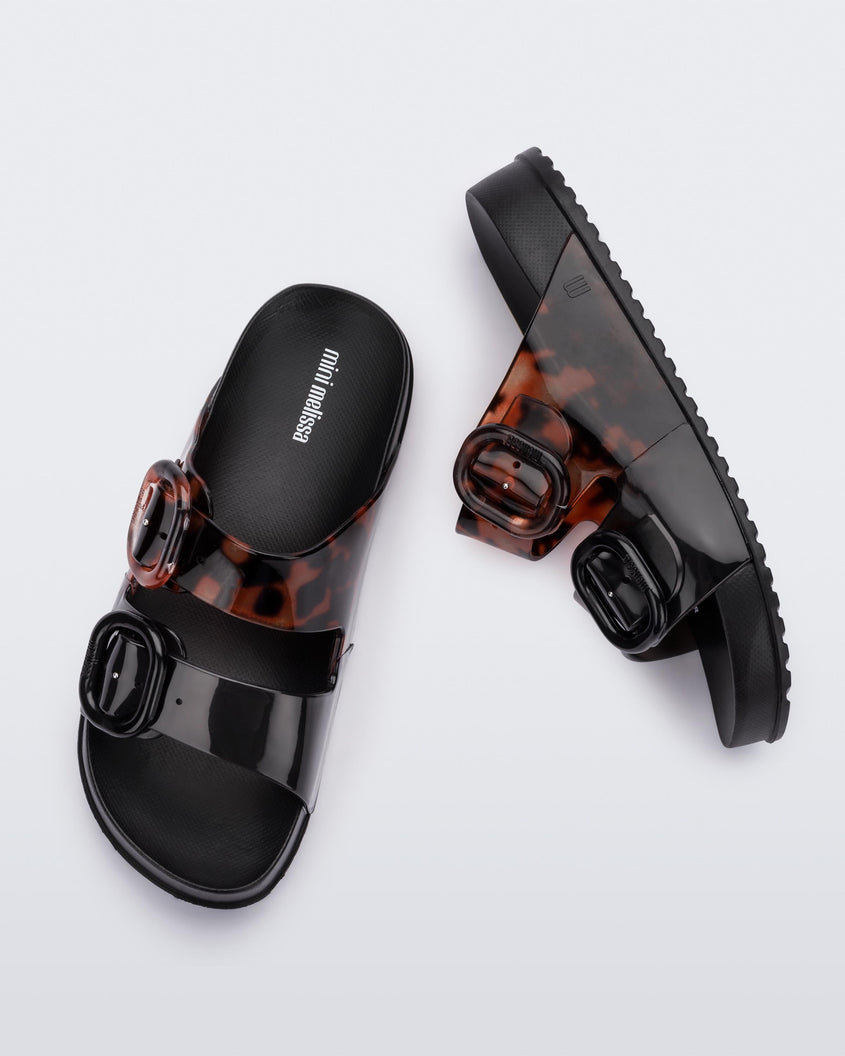 An top and side view of a pair of black tortoiseshell Mini Melissa Cozy slides with two front straps with buckle details