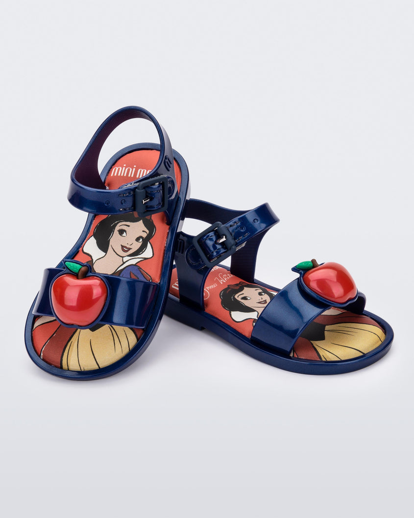 An angled top and side view of a pair of metallic blue Mini Melissa Mar Sandal Princess sandals, leaning on eachother, with an apple detail on the front strap, an ankle strap and Princess Snow White soul