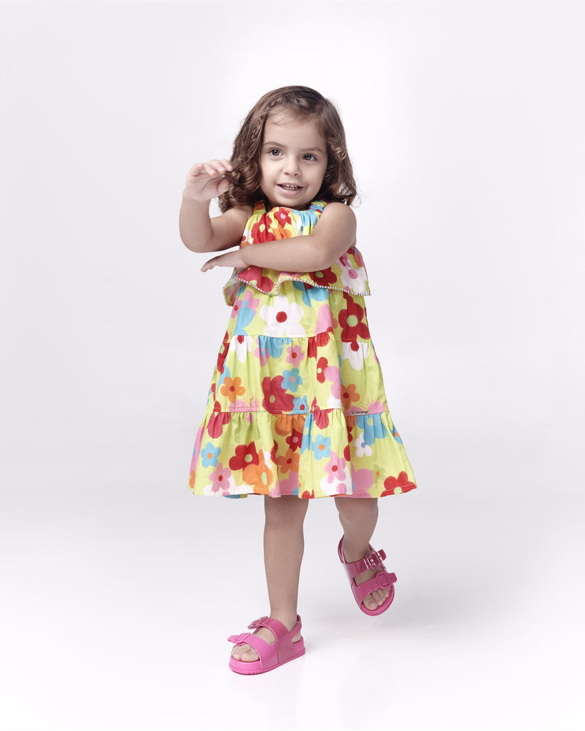 A baby model posing for a picture in a yellow patterned dress and a pair of glitter pink Mini Melissa Cozy sandals with with two front straps with buckle detail