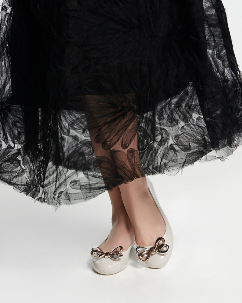 Model's legs in a black skirt wearing a pair of beige Sweet Love Glam ballerina flats with chrome bow