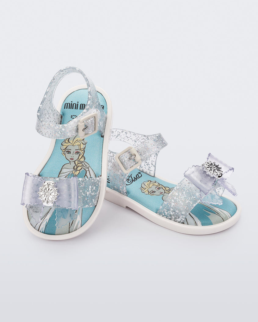 An angled top and side view of a pair of glitter clear Mini Melissa Mar Sandal Princess sandals, leaning on eachother, a snowflake bow detail on the front strap, an ankle strap and Princess Elsa soul