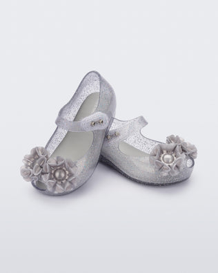 Angled view of a pair of clear grey glitter Ultragirl Springtime baby flat with grey flower and peep toe.