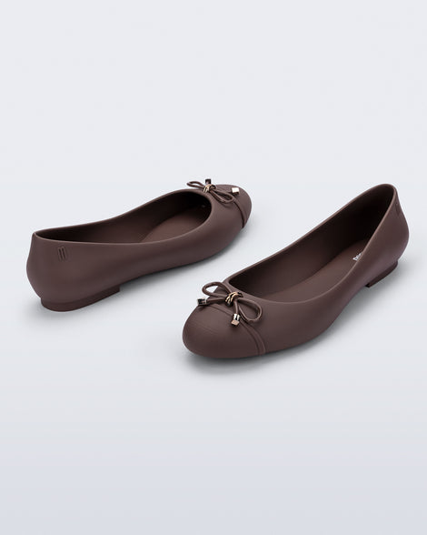 An angled back and side view of a pair of brown Melissa flats with bow detail with gold accents on the bow