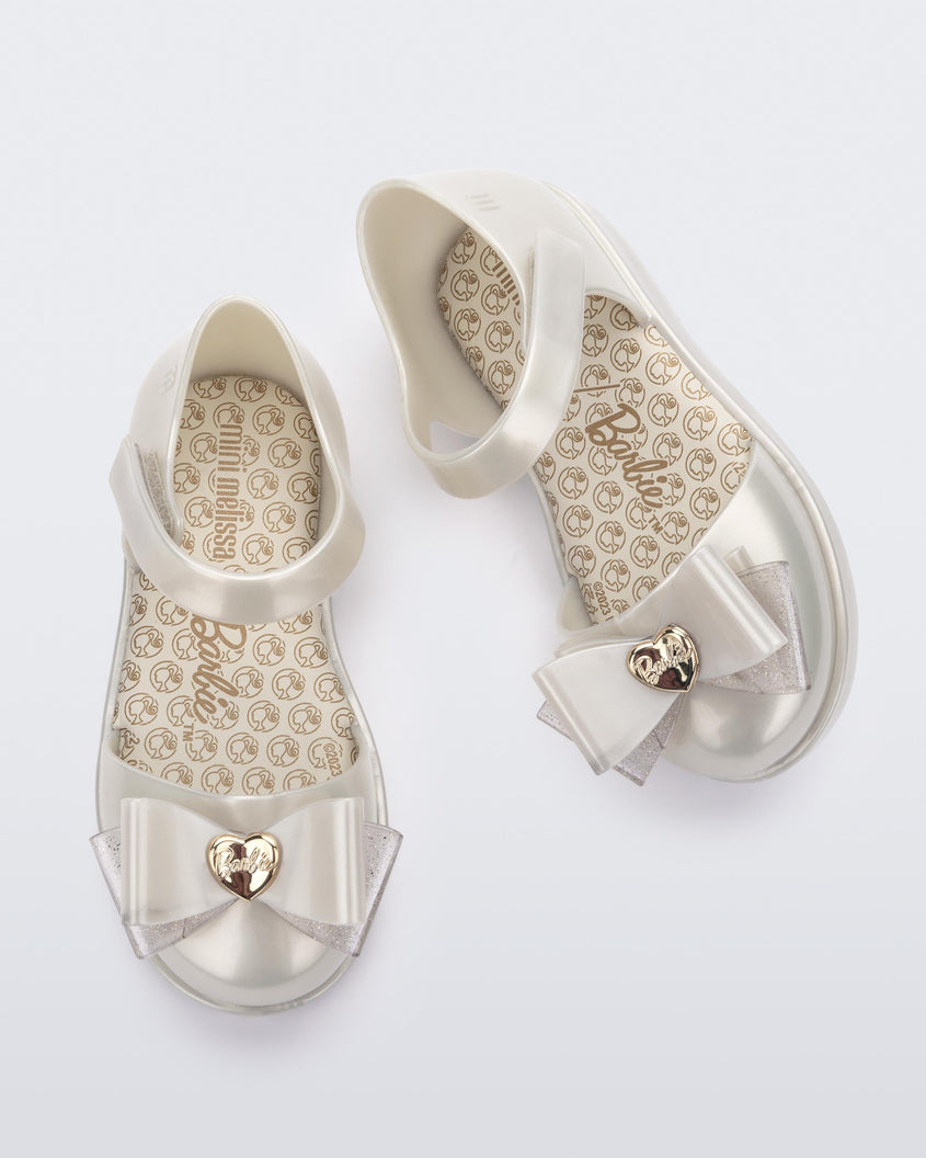 A top view of a pair of white metallic Mini Melissa sandals with a Barbie bow detail on the front toe, white metallic ankle strap and a Barbie logo sole