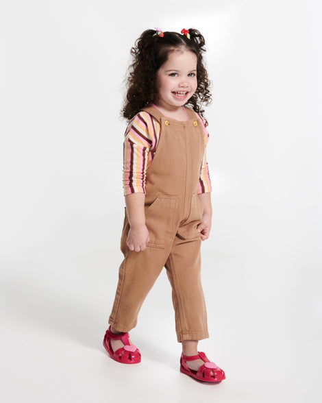 Child model in a brown overalls wearing a pair of red Hip Heart Ballerina baby sandals with pink heart on front