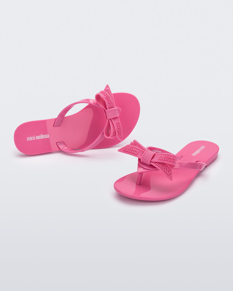 Top and angled view of a pair pink Harmonic M Lover kids flip flops with rainbow Melissa logo on bow