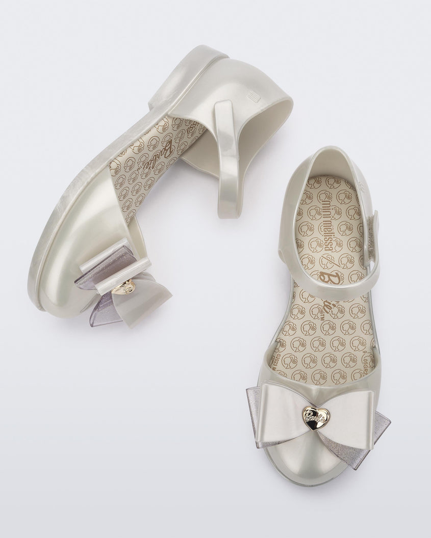 A top and side view of a pair of metallic white Mini Melissa sandals with a Barbie bow detail on the front toe, metallic white ankle strap and a Barbie logo sole