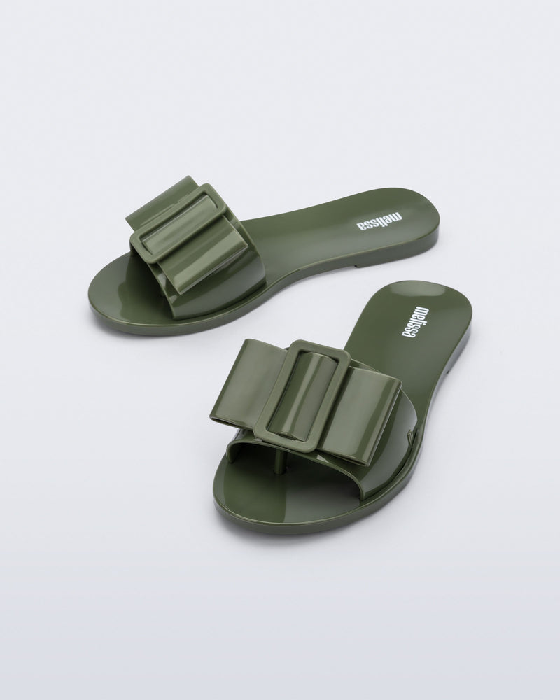 Angled view a pair of dark green Babe slide sandals with large bow on top.