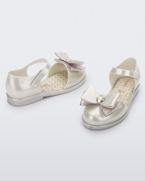 An angled front and side view of a pair of metallic white Mini Melissa sandals with a Barbie bow detail on the front toe, metallic white ankle strap and a Barbie logo sole