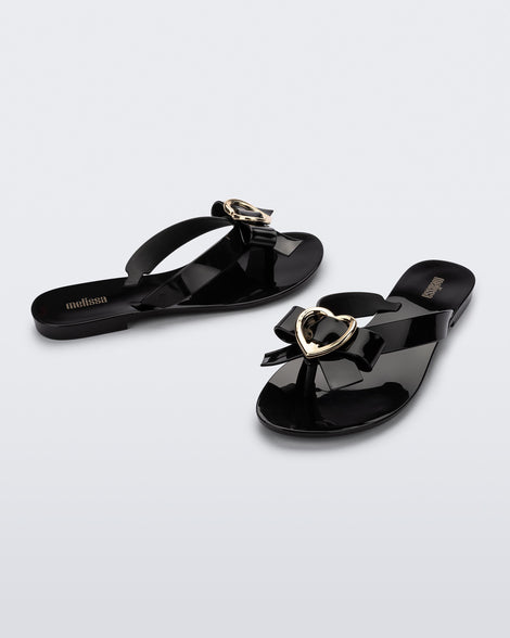 An angled front and side view of a pair of black Melissa Harmonic Heart flip flops with a black bow and gold heart detail on the straps