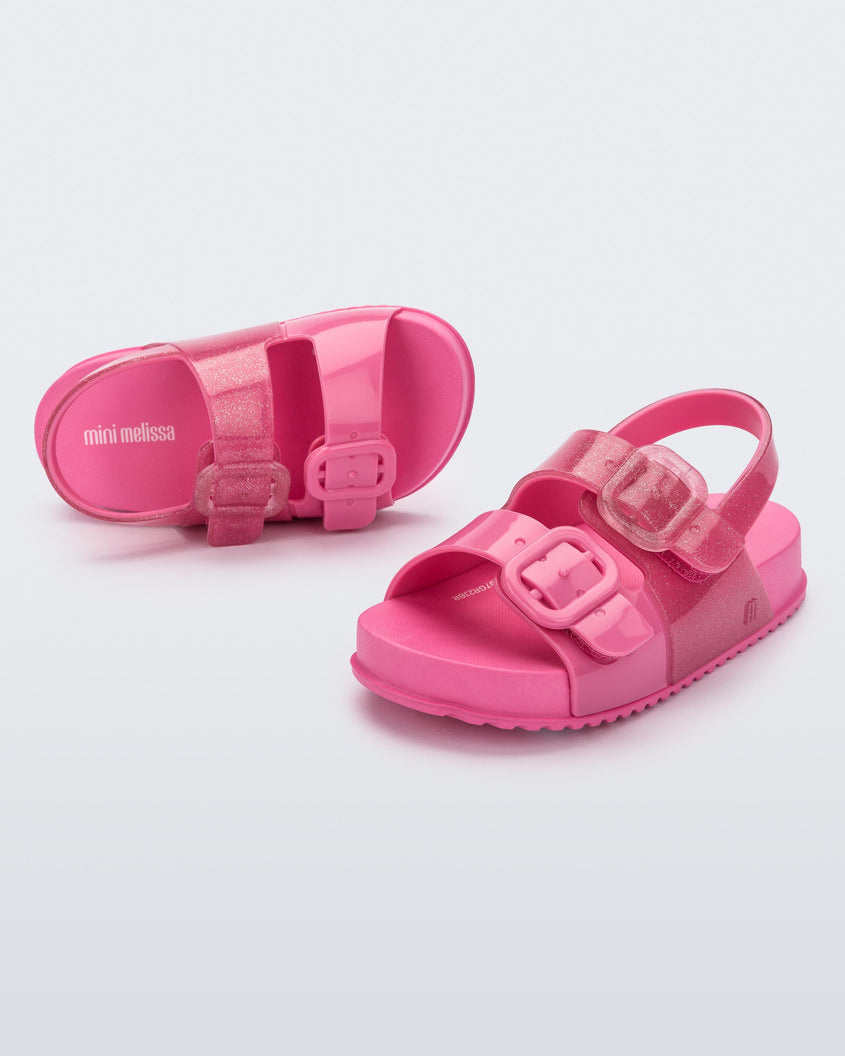 An angled front and top view of a pair of pink glitter Mini Melissa Cozy sandals with two front straps with buckle detail